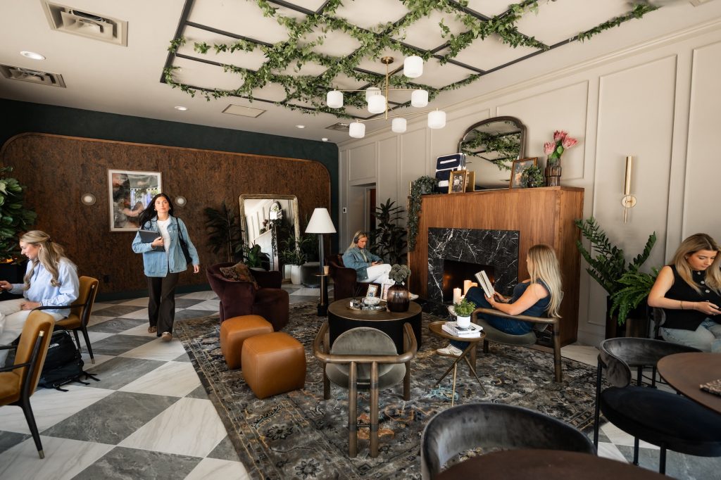 People work and relax in a stylish lounge with plants, elegant furniture, and modern décor—perfect for unwinding after a rambler riverfront tour. Some use laptops or phones, while others chat or walk through the space. The room has a cozy, contemporary feel.