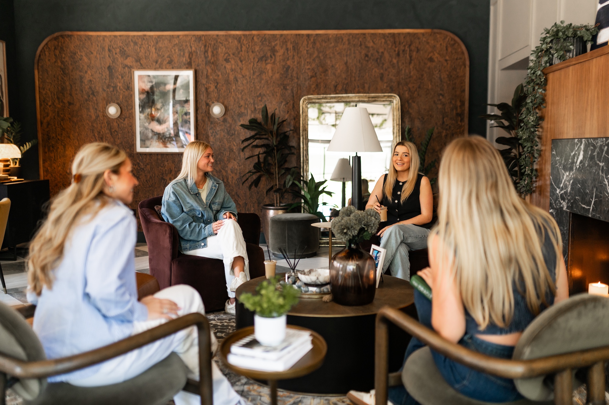 Four women sit in a stylish living room, engaged in conversation after an inspiring rambler riverfront tour. Modern decor, lush plants, and a lit candle create a cozy, inviting atmosphere.