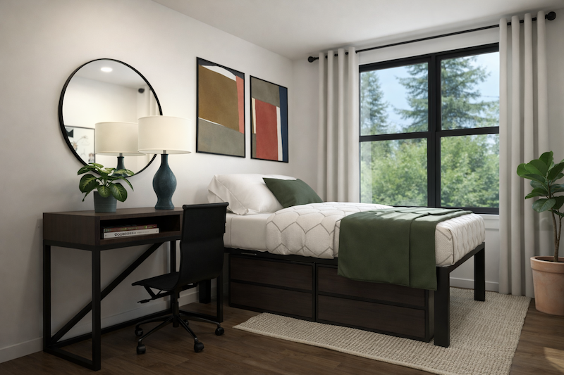 A neatly made bed with green accents sits next to a window, overlooking the Rambler Riverfront; a black desk with a lamp and chair, a round mirror, and modern art on the wall complete the room’s stylish appeal, while a potted plant adds freshness in the corner.