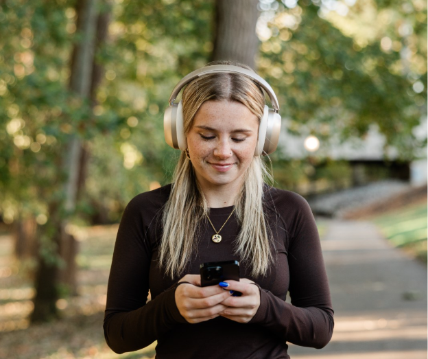 A young woman with long blonde hair wearing over-ear headphones and looking down at a smartphone, perhaps following a Ramber riverfront tour.