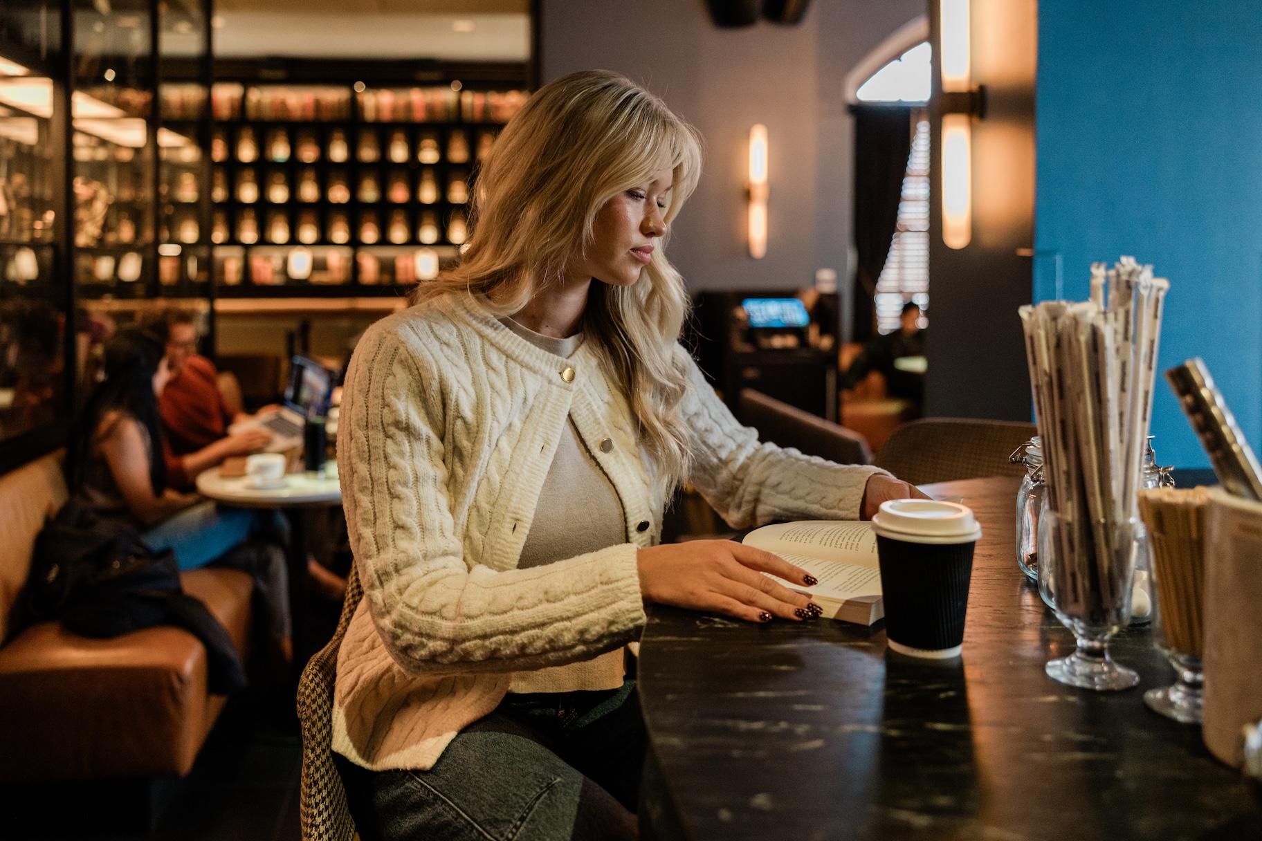 A blonde woman sitting at a dark cafe table reading an open book next to a coffee cup.